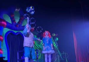 Zdjęcie przedstawia dwóch aktorów na scenie. Jeden tworzy bańki poprzez dmuchanie w rurę.
