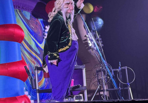 Zdjęcie przedstawia aktora na scenie z uniesionym palcem w górę.