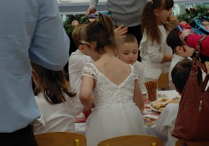 Na zdjęciu widzimy grupkę dzieci, które siedzą wraz z rodzicami przy stole ze słodkim poczęstunkiem.