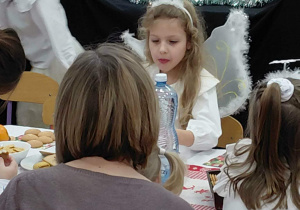 Na zdjęciu widzimy grupkę dzieci, które siedzą wraz z rodzicami przy stole ze słodkim poczęstunkiem.