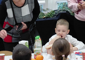 Na zdjęciu widzimy grupkę dzieci, które siedzą wraz z rodzicami przy stole ze słodkim poczęstunkiem.