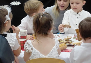 Na zdjęciu widzimy grupkę dzieci, które siedzą wraz z rodzicami przy stole ze słodkim poczęstunkiem.