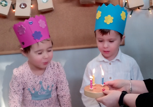 Dzieci siedzą na krzesełkach i zdmuchują świeczki na symbolicznym torcie.
