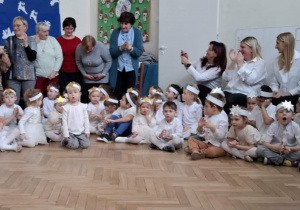 Dzieci w białych strojach ze złotymi gwiazdkami na głosie siedzące na kolędowaniu