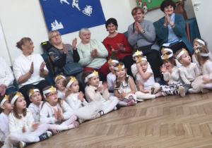 Dzieci w białych strojach ze złotymi gwiazdkami na głosie siedzące na kolędowaniu