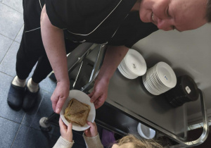 Na zdjęciu widzimy dziewczynkę, która przekazuje talerz z tostem Pani Ani.