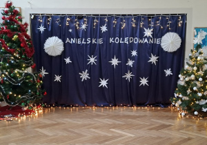 Na zdjęciu widzimy udekorowaną klasę choinkami. Pomiędzy choinkami znajduje się zawieszone granatowy materiał na który powieszony jest biały napis "Anielskie kolędowanie" oraz gdzieniegdzie wiszą białe gwiazdy.