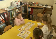 Zdjęcie przedstawia dzieci siedzące przy stoliku podczas zajęć. Na stole rozłożone są obrazki.