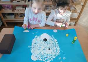 Na zdjęciu widzimy dziewczynki, które przyklejają oczy, noc i uszy na niedźwiedzia polarnego wykonanego stemplując widelec na niebieski karton.