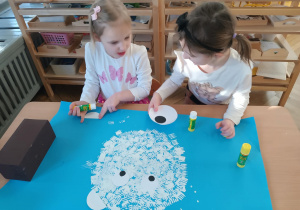 Na zdjęciu widzimy dziewczynki, które przyklejają oczy, noc i uszy na niedźwiedzia polarnego wykonanego stemplując widelec na niebieski karton.