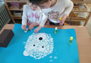 Na zdjęciu widzimy dziewczynki, które przyklejają oczy, noc i uszy na niedźwiedzia polarnego wykonanego stemplując widelec na niebieski karton.