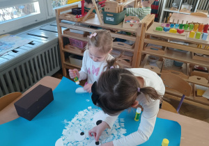 Na zdjęciu widzimy dziewczynki, które przyklejają oczy, noc i uszy na niedźwiedzia polarnego wykonanego stemplując widelec na niebieski karton.