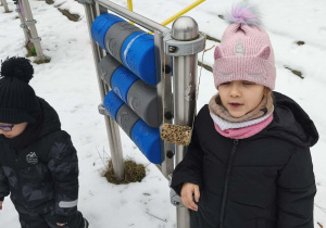 Na zdjęciu widzimy dziewczynkę, która stoi przy zawieszonym karmniku.