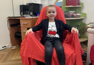 Zdjęcie przedstawia chłopca siedzącego na krześle. Krzesło jest przykryte czerwoną płachtą.