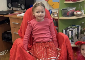 Zdjęcie przedstawia dziewczynkę siedzącą na krześle. Krzesło jest przykryte czerwoną płachtą.