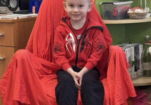 Zdjęcie przedstawia chłopca siedzącego na krześle. Krzesło jest przykryte czerwoną płachtą.