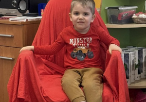 Zdjęcie przedstawia chłopca siedzącego na krześle. Krzesło jest przykryte czerwoną płachtą.