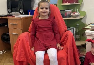 Zdjęcie przedstawia dziewczynkę siedzącą na krześle. Krzesło jest przykryte czerwoną płachtą.