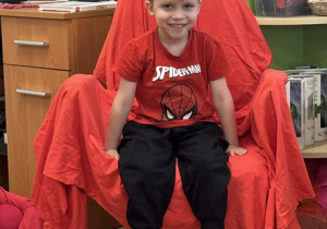 Zdjęcie przedstawia chłopca siedzącego na krześle. Krzesło jest przykryte czerwoną płachtą.