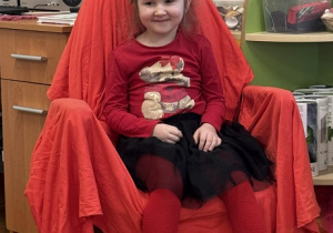 Zdjęcie przedstawia dziewczynkę siedzącą na krześle. Krzesło jest przykryte czerwoną płachtą.