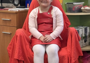 Zdjęcie przedstawia dziewczynkę siedzącą na krześle. Krzesło jest przykryte czerwoną płachtą.