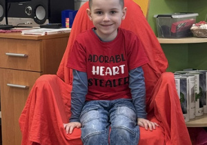 Zdjęcie przedstawia chłopca siedzącego na krześle. Krzesło jest przykryte czerwoną płachtą.