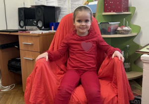 Zdjęcie przedstawia dziewczynkę siedzącą na krześle. Krzesło jest przykryte czerwoną płachtą.