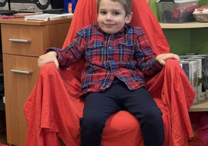 Zdjęcie przedstawia chłopca siedzącego na krześle. Krzesło jest przykryte czerwoną płachtą.