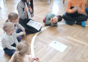 Dzieci siedzą w kole i pani pokazuje im ilustracje.