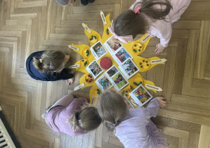 Zdjęcie przedstawia dzieci oglądające zdjęcia rozłożone na słoneczku urodzinowym.