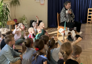 Zdjęcie przedstawia dzieci siedzące na podłodze, podczas rozmowy z mężczyzną. Dzieci podnoszą rękę do góry, by zgłosić się do odpowiedzi.