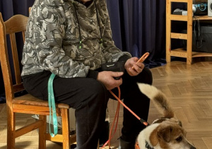 Zdjęcie przedstawia mężczyznę siedzącego na krześle. Mężczyzna trzyma psa na smyczy.