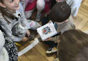 Na zdjęciu widzimy dzieci, które oglądają książkę i jej okładkę.