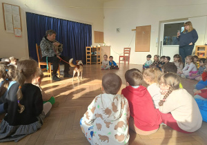 Zdjęcie przedstawia dzieci siedzące w półkole. Przed nimi siedzi mężczyzną z dwójką zwierząt- psem i kotem.