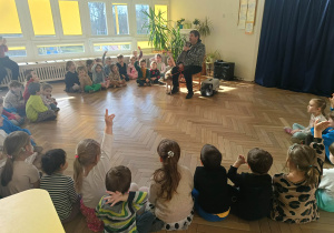 Zdjęcie przedstawia dzieci siedzące w półkole. Przed nimi siedzi mężczyzną z dwójką zwierząt- psem i kotem. Dzieci zgłaszają się do odpowiedzi.