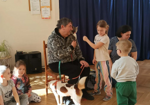 Zdjęcie przedstawia mężczyznę trzymającego kota, obok stoi pies. Do mężczyzny podeszły dzieci, aby pogłaskać zwierzęta.