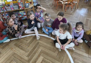 Na zdjęciu widzimy dzieci, które siedzą na podłodze w parach i pokazują, że kaszlą pod wpływem "wąchania" dymu papierosowego.