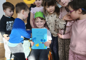 Na zdjęciu widzimy Jubilata w koronie i z upominkiem od dzieci. Wokół Jubilata znajdują się dzieci.