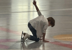 Zdjęcie przedstawia chłopca na lodzie, rozpoczynającego swój występ. Chłopiec klęczy na lodzie, z ręką wyciągniętą do góry.