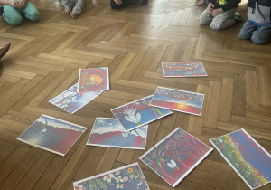 Zdjęcie przedstawia dzieci siedzące wokół rozłożonych kartek z ilustracjami.
