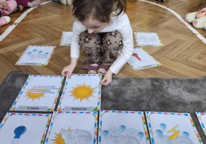 Na zdjęciu widzimy dziewczynkę, która kładzie na dywanik ilustrację przedstawiającą słońce.