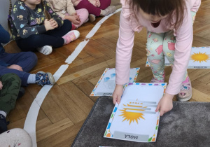 Na zdjęciu widzimy dziewczynkę, która kładzie na dywanik ilustrację przedstawiającą mgłę.