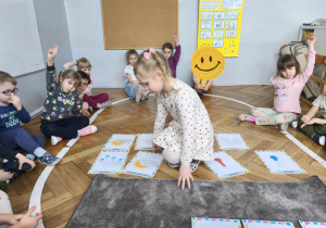 Na zdjęciu widzimy dziewczynkę, która bierze z podłogi ilustrację przedstawiającą zachmurzenie.