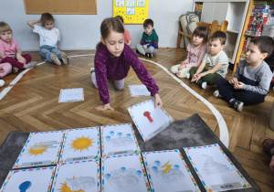 Na zdjęciu widzimy dziewczynkę, która kładzie na dywanik ilustrację przedstawiającą zimno.