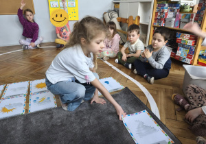 Na zdjęciu widzimy dziewczynkę, która kładzie na dywanik ilustrację przedstawiającą wiatr.