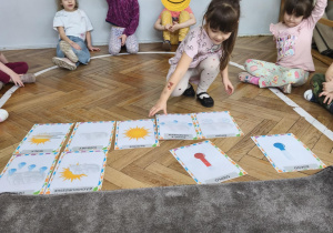 Na zdjęciu widzimy dziewczynkę, która podnosi z podłogi ilustrację przedstawiającą śnieg z deszczem.