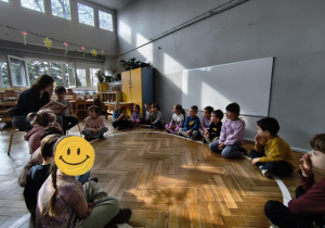 Na zdjęciu widzimy grupkę dzieci, które słuchają czytanej przez mamę Tymona książki.