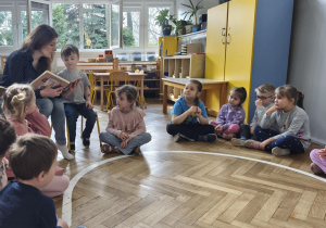 Na zdjęciu widzimy grupkę dzieci, które słuchają czytanej przez mamę Tymona książki.