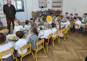 Zdjęcie przedstawia dzieci siedzące przy stole oraz pracownika przedszkola, który stoi obok.