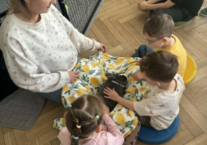 Zdjęcie przedstawia kobietę i dzieci przykrytych kocem. Na kocu leży królik, którego głaszczą dzieci.